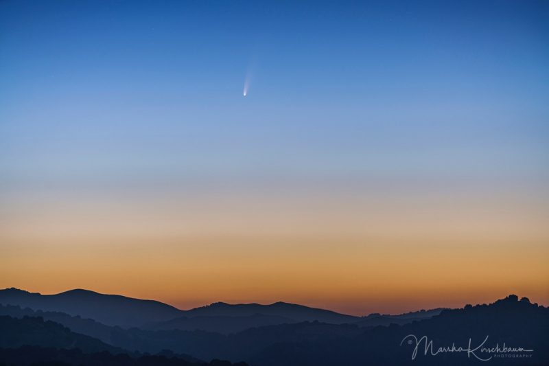 A comet with a split tail set against a bright twilight sky over blue hills.