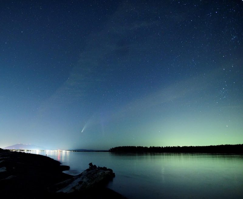 Comet and aurora against a starry medium blue sky, over a body of water.