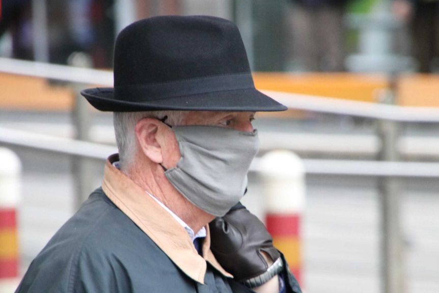 A man with grey hair wearing a hat, mask and gloves walks down a street.
