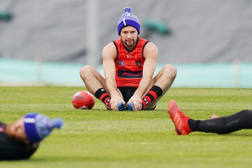 A football player wearing a beanie sits on the ground during training with hands touching his feet.