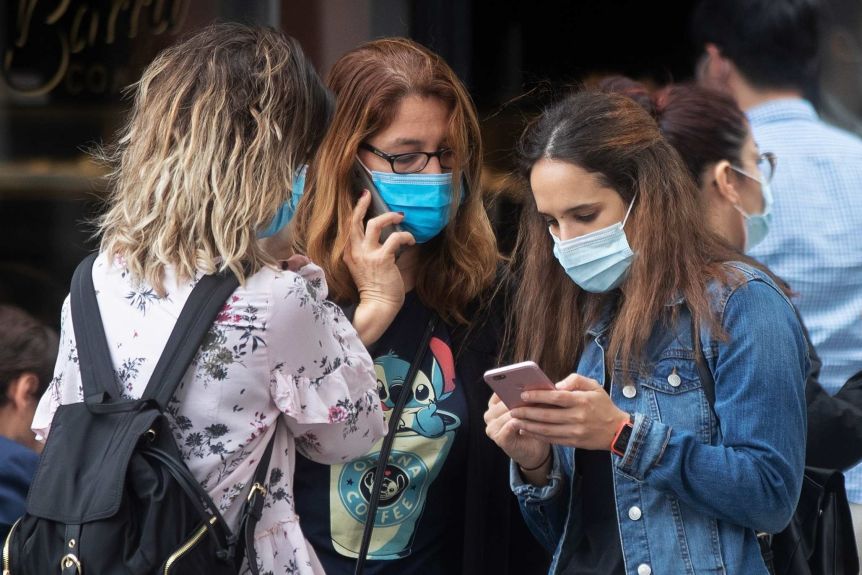 Three young women wearing masks, stand together, and use their phones